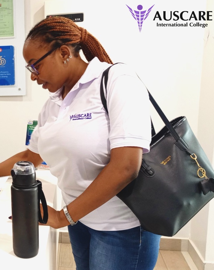 Woman in a white AUSCARE polo at a reception, holding a black water bottle with a black handbag over her shoulder; AUSCARE logo visible in background.