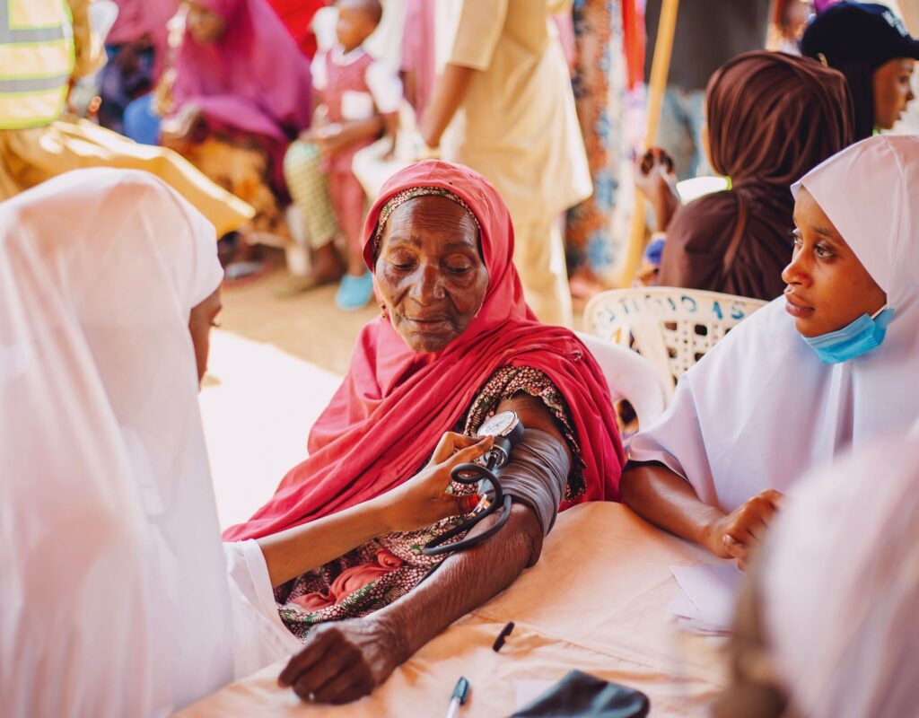 Nurse uses a stethoscope to measure an elderly woman's blood pressure at a busy clinic in the background note of other people present.