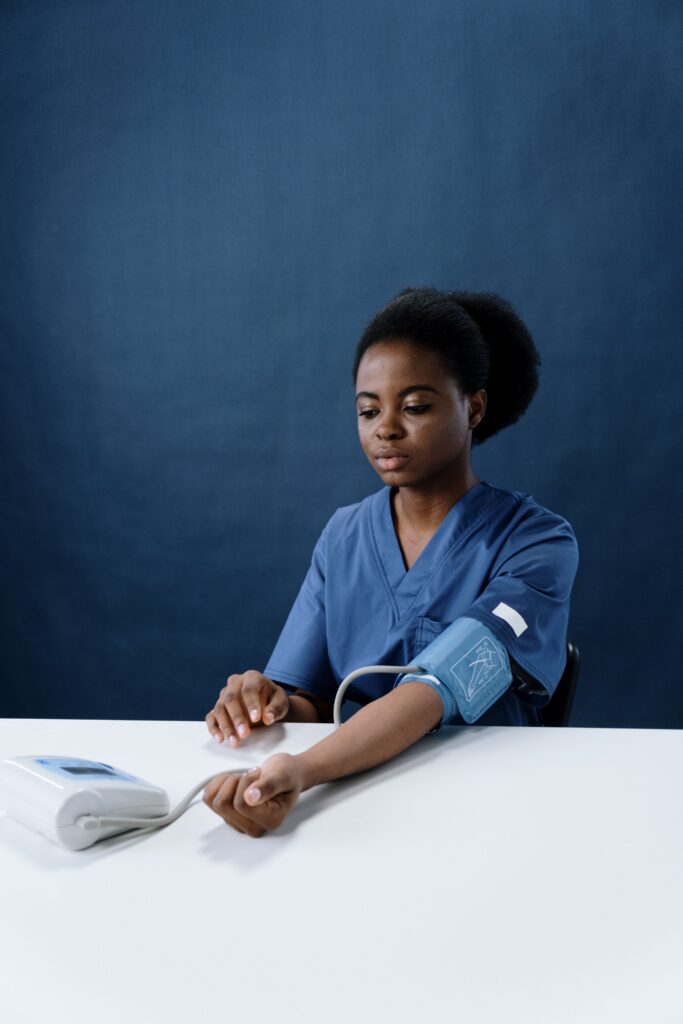 Black woman in blue scrubs seated at a white table, applying a blood pressure cuff to her arm beside a monitor.