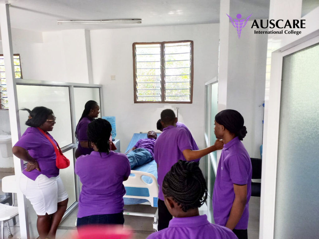 Nursing students in purple shirts gather around a patient lying on a hospital bed in a bright clinical room. AUSCARE logo visible in the top right corner.