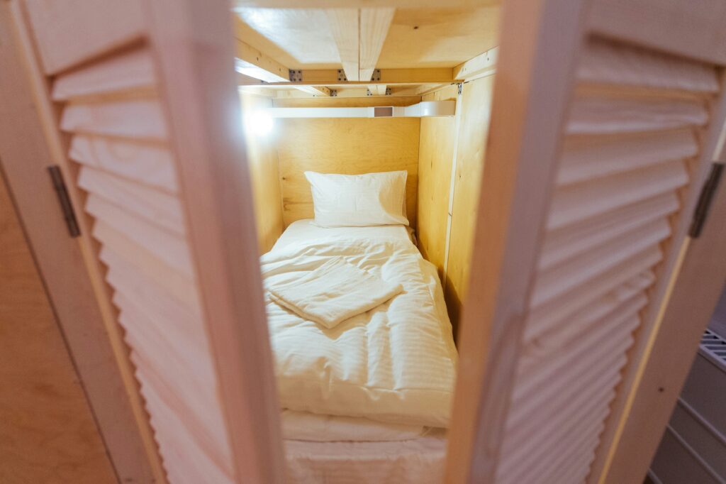 Inside a small wooden bunk-bed alcove with white bedding, viewed through door slats with soft lighting
