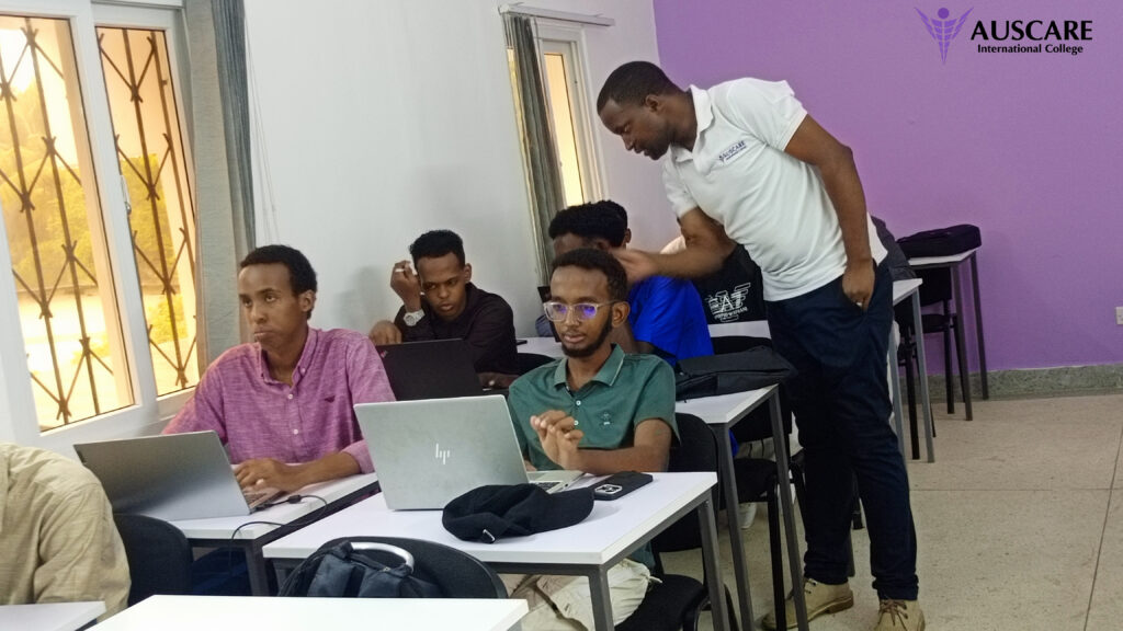 Group of students at desks using laptops in a classroom while a standing instructor assists a student; purple wall with AUSCARE logo in the corner.