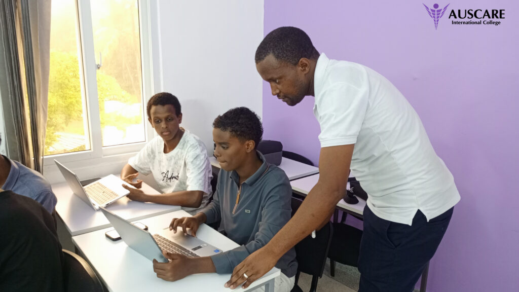 Teacher assisting two students at laptops in a bright classroom with a purple wall and AUSCARE logo in the corner.