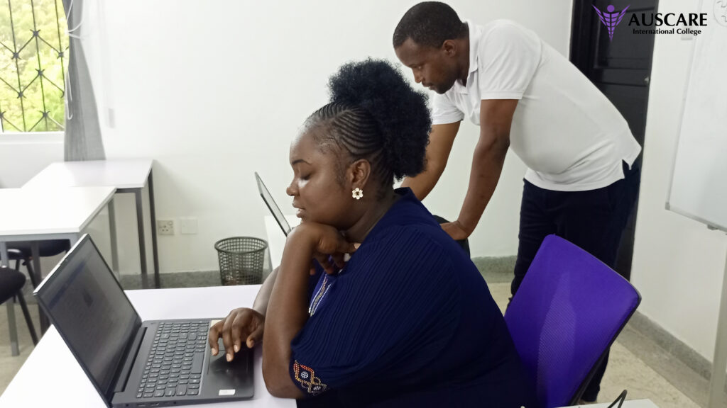 Woman seated at a desk typing on a laptop while a man stands behind her offering help; AUSCARE logo in the top-right corner.