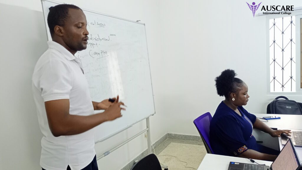 Instructor speaking and writing on a whiteboard in a classroom, with a student at a laptop and AUSCARE logo in the corner.