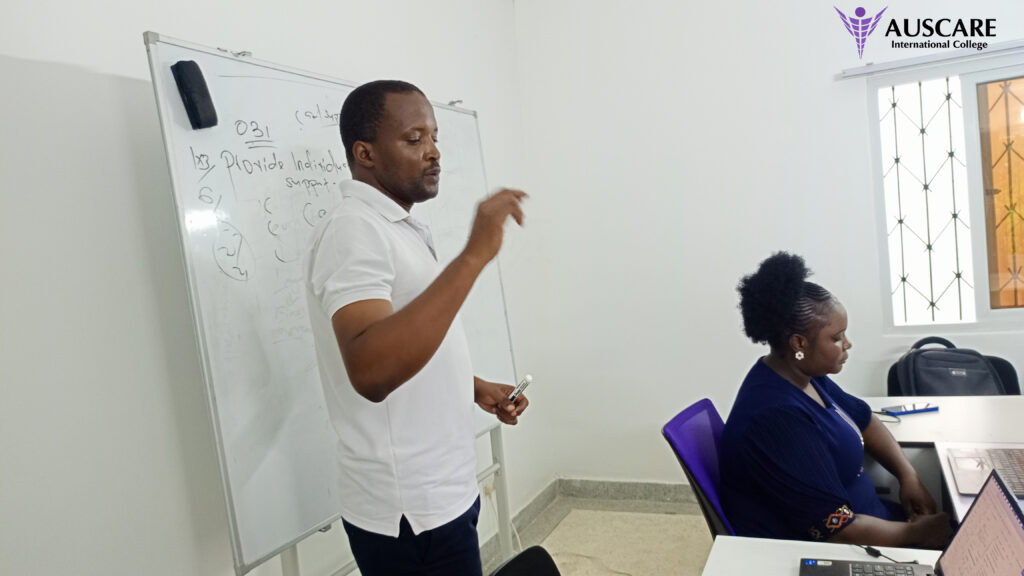 Male instructor writes on a whiteboard at the front of a classroom as a student watches; AUSCARE International College logo appears in the top right corner.