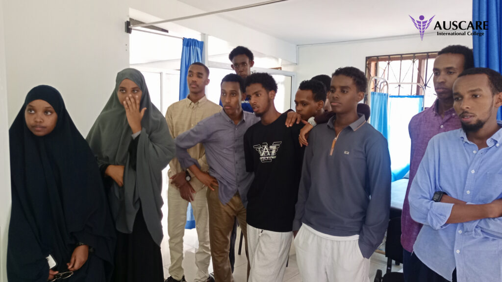 Group of diverse students indoors, two women in head coverings at the left, others in casual clothes, standing together in a classroom-like setting with AUSCARE logo in the corner