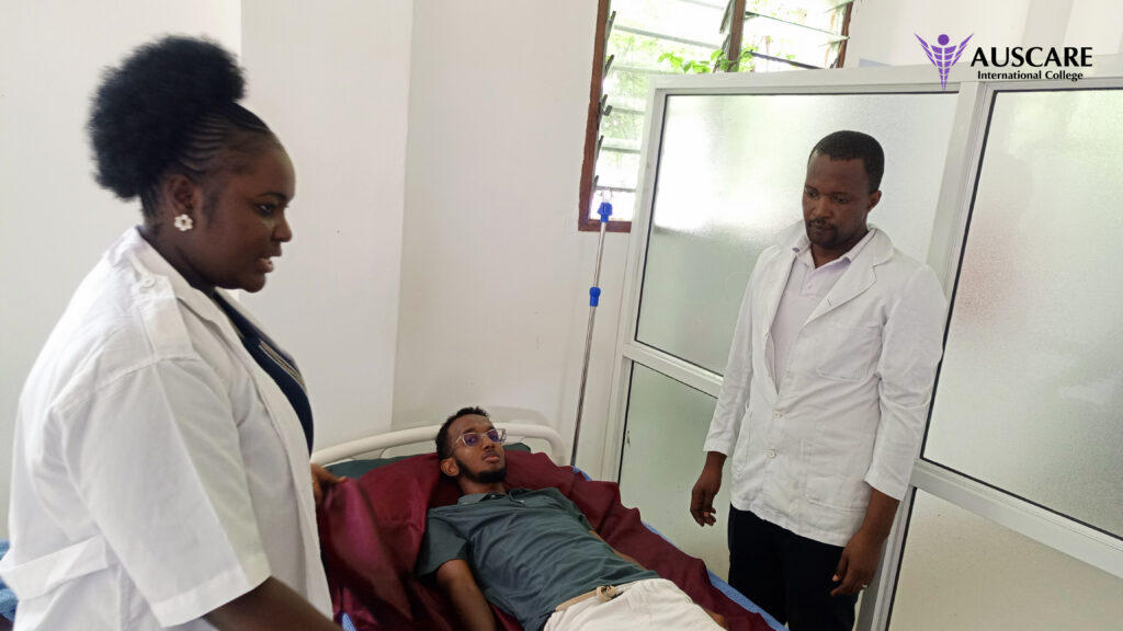 Male patient lying in a hospital bed with red blanket, two clinicians in white coats stand nearby in a clinical room; IV stand visible, AUSCARE logo shows in the corner.